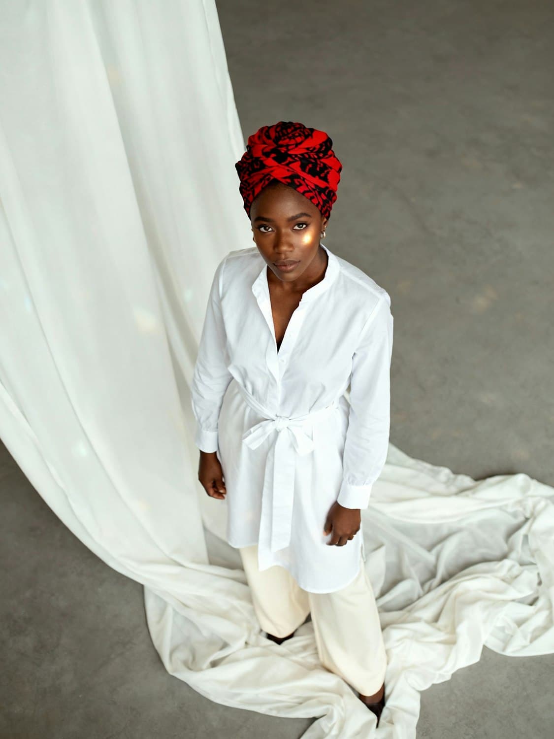 A woman in a red patterned headwrap and white tunic standing beside flowing white fabric, looking up calmly