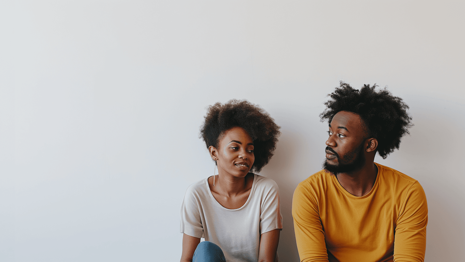 A couple sitting against a pale wall, turned toward each other mid-conversation